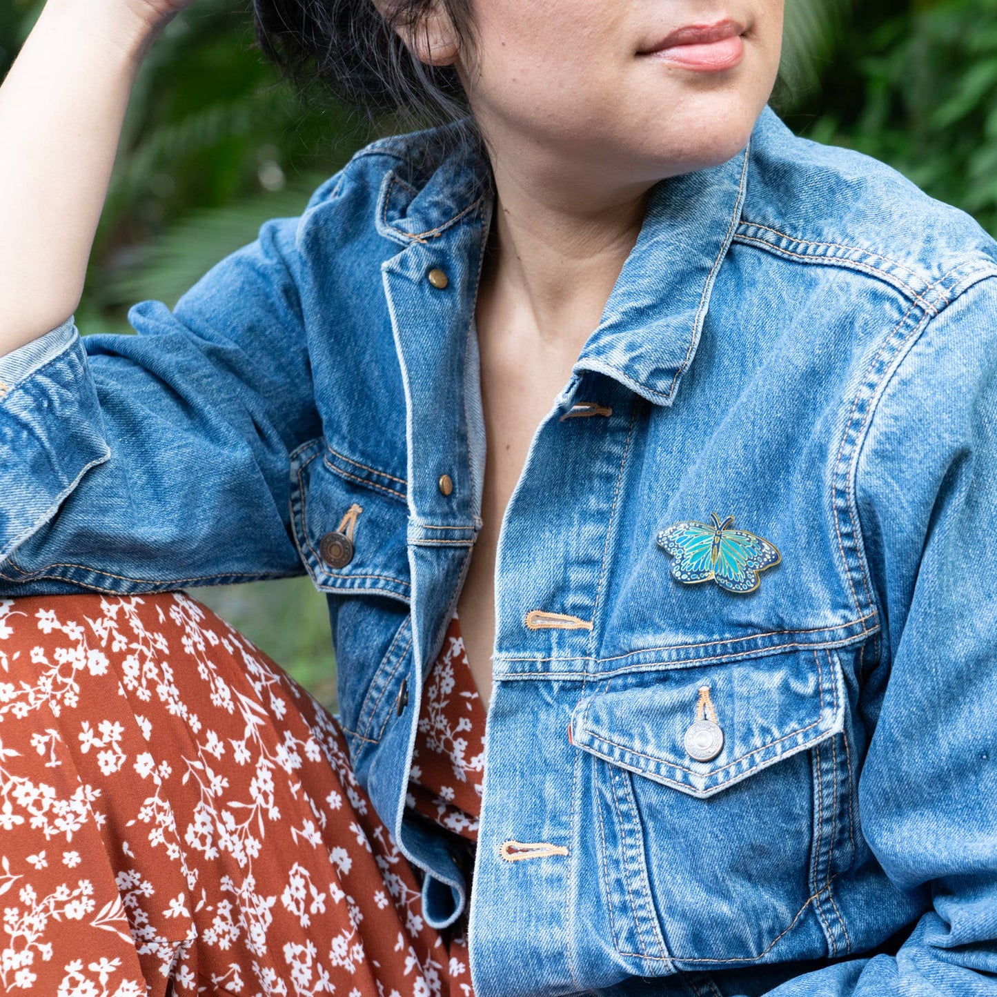 Person wearing a denim jacket with a butterfly pin, standing outdoors.